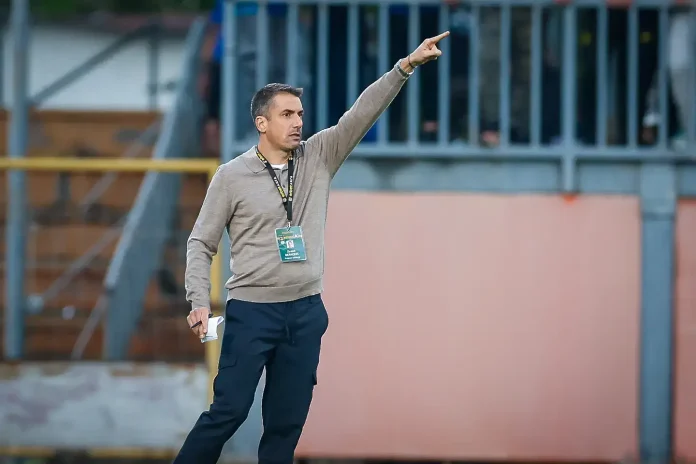 Sports coach on a field, pointing with his right arm as he gives directions, badge hanging from a lanyard around his neck