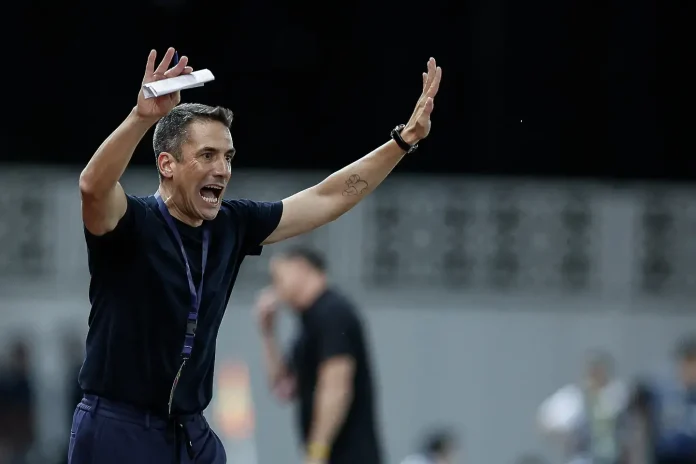 Coach celebrating on a sports field with arms raised, holding a folded paper, wearing a dark shirt and a lanyard.