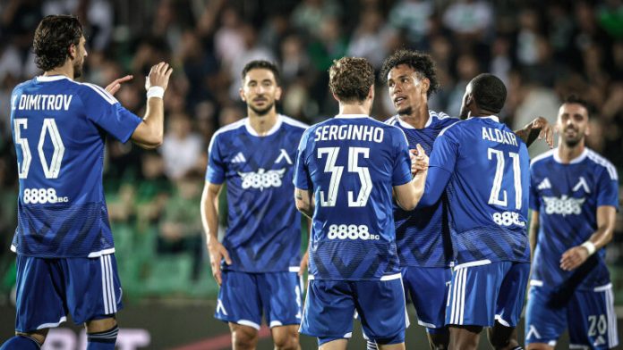 Soccer players in blue uniforms celebrate together on a field after a goal, with arms raised and smiling faces, a crowded stadium in the background.