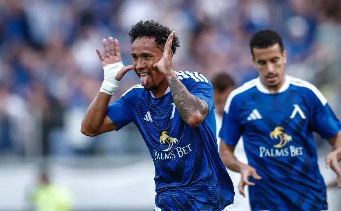 Soccer player in blue kit celebrating with hands by ears, audience blurred in stadium behind him.