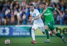 Веласкес получава още една опция в защита Two soccer players chase the ball on a green field as fans watch from the stands, one in white and blue and the other in green.