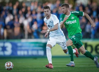 Веласкес получава още една опция в защита Two soccer players chase the ball on a green field as fans watch from the stands, one in white and blue and the other in green.