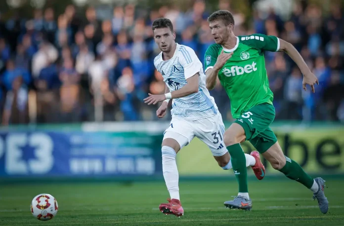 Two soccer players chase the ball on a green field as fans watch from the stands, one in white and blue and the other in green.
