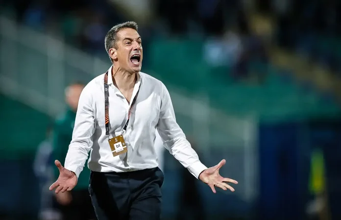 Coach in a white shirt shouts with arms outstretched on a sports field, wearing a lanyard and pass attached to it.