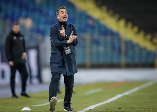 Football coach on a sideline gesturing emphatically during a match in a stadium, wearing a dark coat and lanyard.