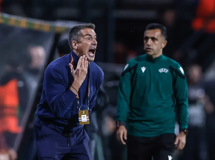 Coach in blue screams with hand to face on the sideline, badge hanging from lanyard visible on chest level.