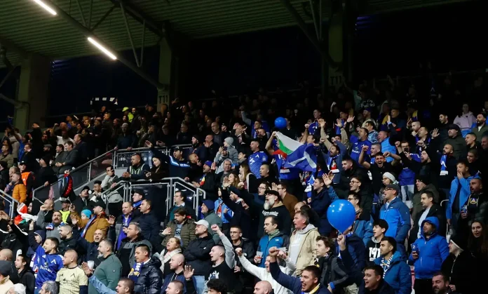 Crowd of sports fans in a stadium stands, many wearing blue, waving flags and blue balloons.