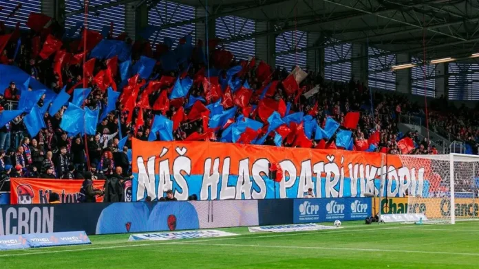 Supporters in a stadium waving red and blue flags behind a large orange banner with white letters.