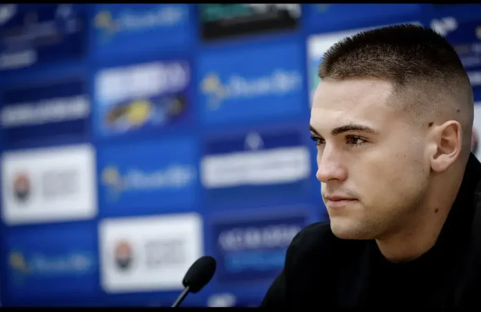 Portrait of a young man with short hair speaking at a press conference, microphone in foreground and a blurred blue backdrop behind him.