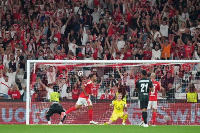 Soccer goal area: a red-team attacker shoots as the yellow-clad goalkeeper reacts on the ground, with a cheering crowd behind the net.