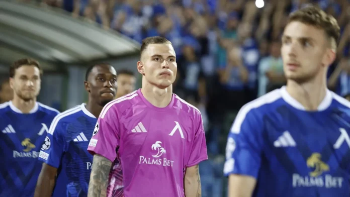 Soccer players walk onto the field before kickoff; a goalkeeper in pink stands center with teammates in blue nearby.