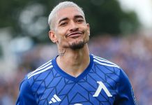 Уелтон – статистика, клубове и роля Soccer player with short platinum hair wearing a blue geometric-patterned jersey, smirking toward the camera on the field.