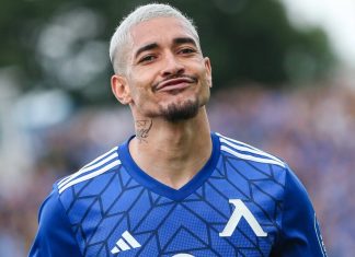 Уелтон – статистика, клубове и роля Soccer player with short platinum hair wearing a blue geometric-patterned jersey, smirking toward the camera on the field.