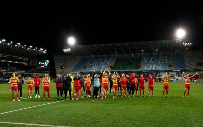 Soccer team in red and yellow uniforms stands on a lit pitch after a match, waving to fans in a night stadium.