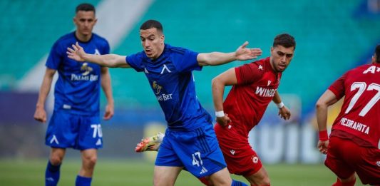 Задава се ново голямо дерби Левски – ЦСКА Soccer players in blue and red jerseys chase a ball on a green field during a match with a teal seating backdrop.