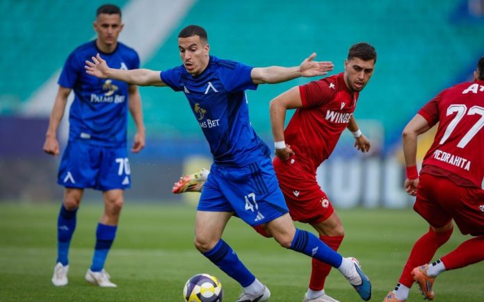 Soccer players in blue and red jerseys chase a ball on a green field during a match with a teal seating backdrop.