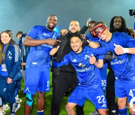Group of soccer players in blue uniforms celebrate and pose for a photo on a green field after a win, smiling and giving thumbs up to the camera.