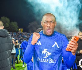 Football player in a blue jersey celebrates on the field with a clenched fist and open mouth as smoke rises behind him.