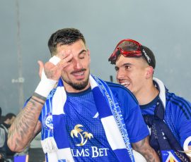 Two teammates in blue uniforms celebrate together, one wiping his eye with a white towel as the other grins beside him in a stadium.