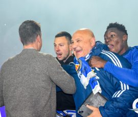Group of men in blue athletic jackets celebrate and hug in a stadium setting, one man in gray sweater facing away.