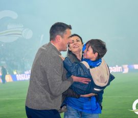 Family on a sports field: man holding a smiling child while a woman watches, all embracing in a stadium with a blue smoke-filled backdrop and banners in the background.