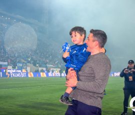 Man lifts a smiling boy in a blue jersey on a soccer field, with a cheering stadium crowd in the background.