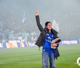 Woman on a sports field raises her fist in celebration, wearing a blue Palms Bet jersey and a dark jacket.