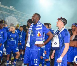 Soccer players in blue kits celebrate on the field with scarves, smiling as fans cheer in a packed stadium behind them.