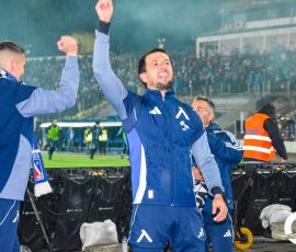 Athlete in a blue tracksuit raises a fist in celebration on a rainy stadium sideline with teammates and a cheering crowd behind him.