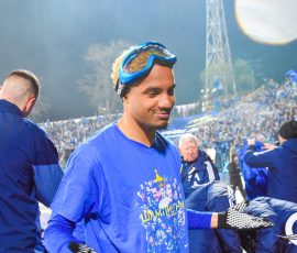 Athlete in blue jersey with swim goggles on his head, smiling, stadium crowd in blue behind him.