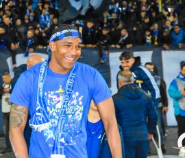 Smiling athlete in a blue shirt and headband, with fans cheering in blue in the background during a celebration event.