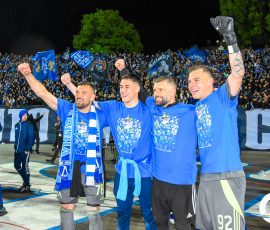 Four soccer players in blue shirts celebrate with raised arms as a rain-soaked stadium full of cheering fans waves blue banners in the background.