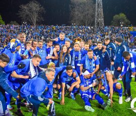 A large group of blue-clad soccer players celebrate on the field amid a cheering crowd under stadium lights in the rain at night.