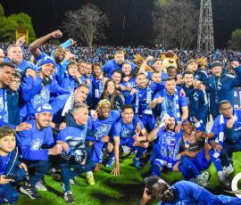 Group of blue-clad soccer players celebrating together on the field with a cheering crowd in the stands behind them under stadium lights on a rainy night.