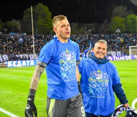 Two soccer players in blue jerseys celebrate on a rain-soaked field with a packed stadium in the background.