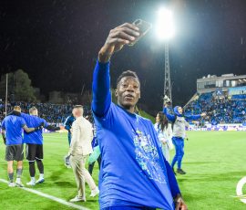 Soccer player in blue raises a phone for a selfie amid celebration on a floodlit field.