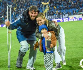 Smiling man with crutches stands with a woman and child on a soccer field, with a cheering crowd in the stands behind them.