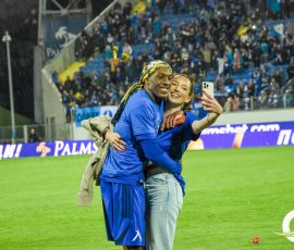 Two athletes in blue hug and selfie on a rain-lit soccer field with a crowded stadium backdrop.