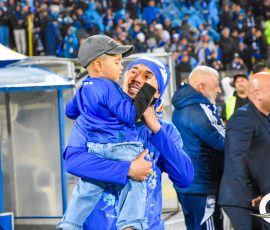Man in blue jacket lifts smiling child wearing a gray cap near the sideline of a stadium with a cheering crowd behind them