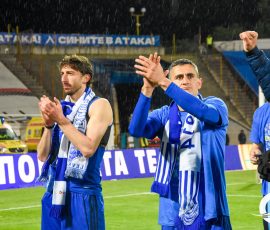 Soccer players in blue uniforms celebrate on a rain-soaked field, fists raised and clapping with banners visible in the stands behind them