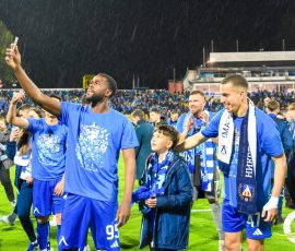 Soccer players in blue uniforms celebrate on a rain-soaked field with a cheering crowd in the stands behind them.