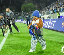 Young child in a blue beanie runs on a soccer field as photographers crouch nearby in the rain, crowd in the stands behind.