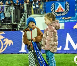 Two young children in winter coats stand on a grassy soccer field, holding blue and white scarves as stadium spectators watch behind a blue banner.