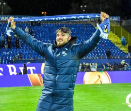 Man in a blue puffer jacket raises a blue scarf above his head on a soccer pitch at night stadium grounds.