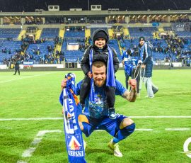 Man kneeling on a soccer field with a smiling child on his shoulders, blue team scarves visible, stands full of fans in the background.