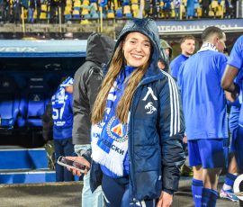Smiling woman in a blue rain jacket and scarf stands on a stadium sideline, holding a smartphone against a background of fans in blue and yellow.
