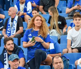 Woman with long curly hair in a blue jersey sits in stadium seats, holding a green smartphone.