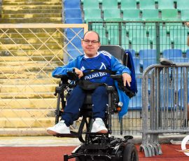 Smiling man in a blue sports jersey sits in a wheelchair on the track beside a metal barrier in a stadium. Bleachers are in the background.