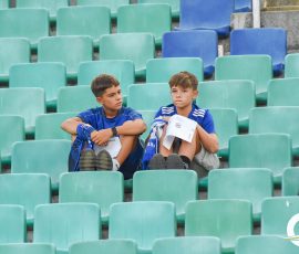 Two boys in blue shirts sit among teal stadium seats, looking sideways and holding papers or booklets.
