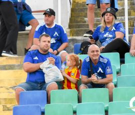 Spectators in matching blue jerseys sit in stadium seats, with a man eating popcorn and a young girl in a yellow-red jersey nearby